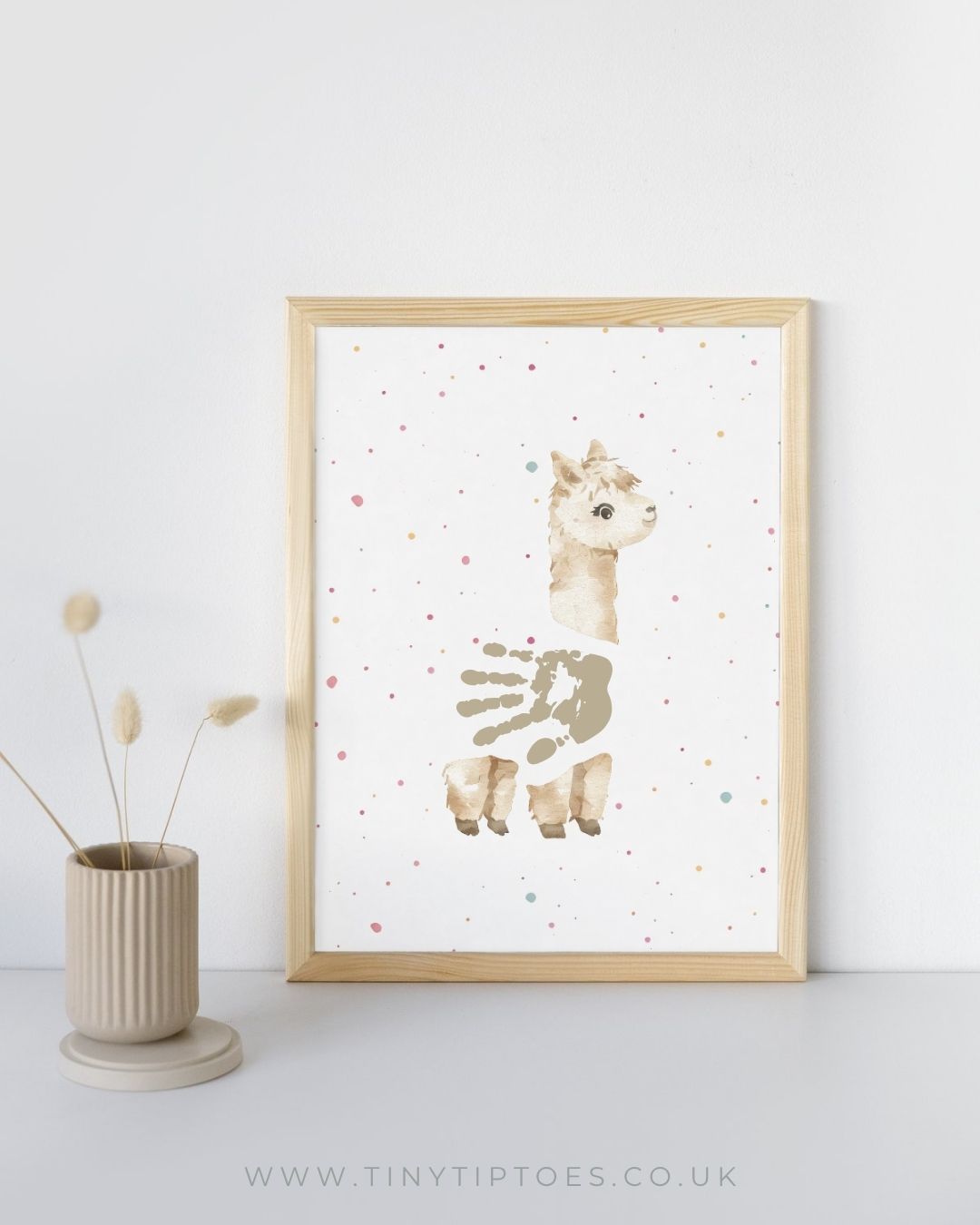 Framed artwork of an alpaca with a handprint body on a white background with colorful dots, next to a vase with dried plants.