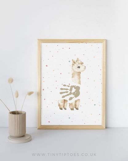 Framed artwork of an alpaca with a handprint body on a white background with colorful dots, next to a vase with dried plants.