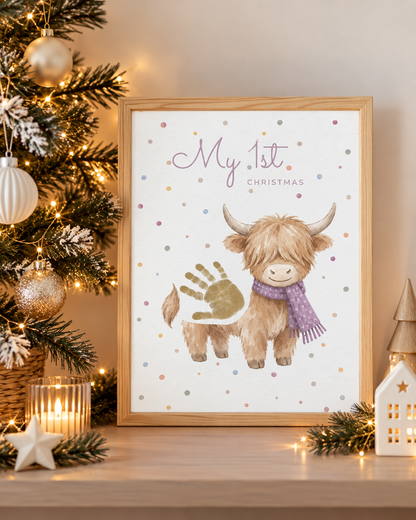 A framed personalized Christmas art print featuring a watercolor Highland cow with a purple scarf and a gold handprint, next to a decorated Christmas tree on a wooden shelf.