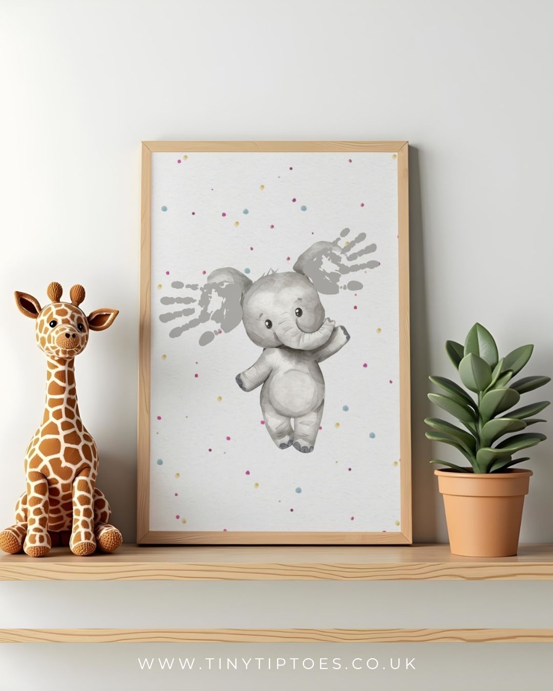 Framed children's art print of an elephant with handprint ears, sitting on a shelf with a toy giraffe and plant.