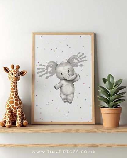 Framed children's art print of an elephant with handprint ears, sitting on a shelf with a toy giraffe and plant.