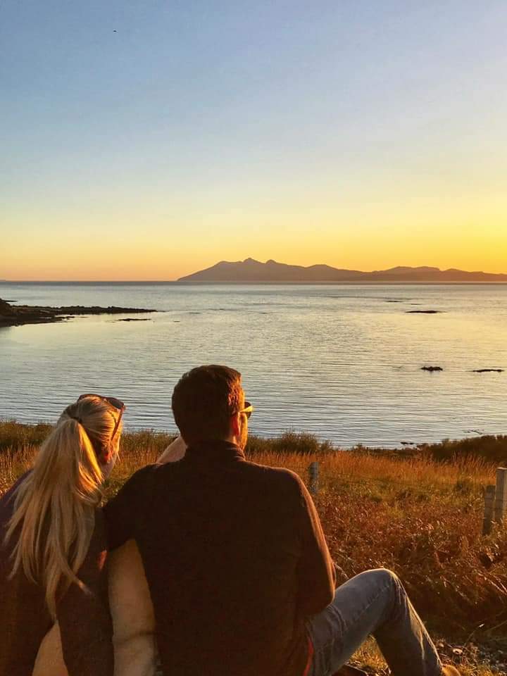 Husband and wife team, Michael and Emma with their dog looking at mountains, sea and sunset.