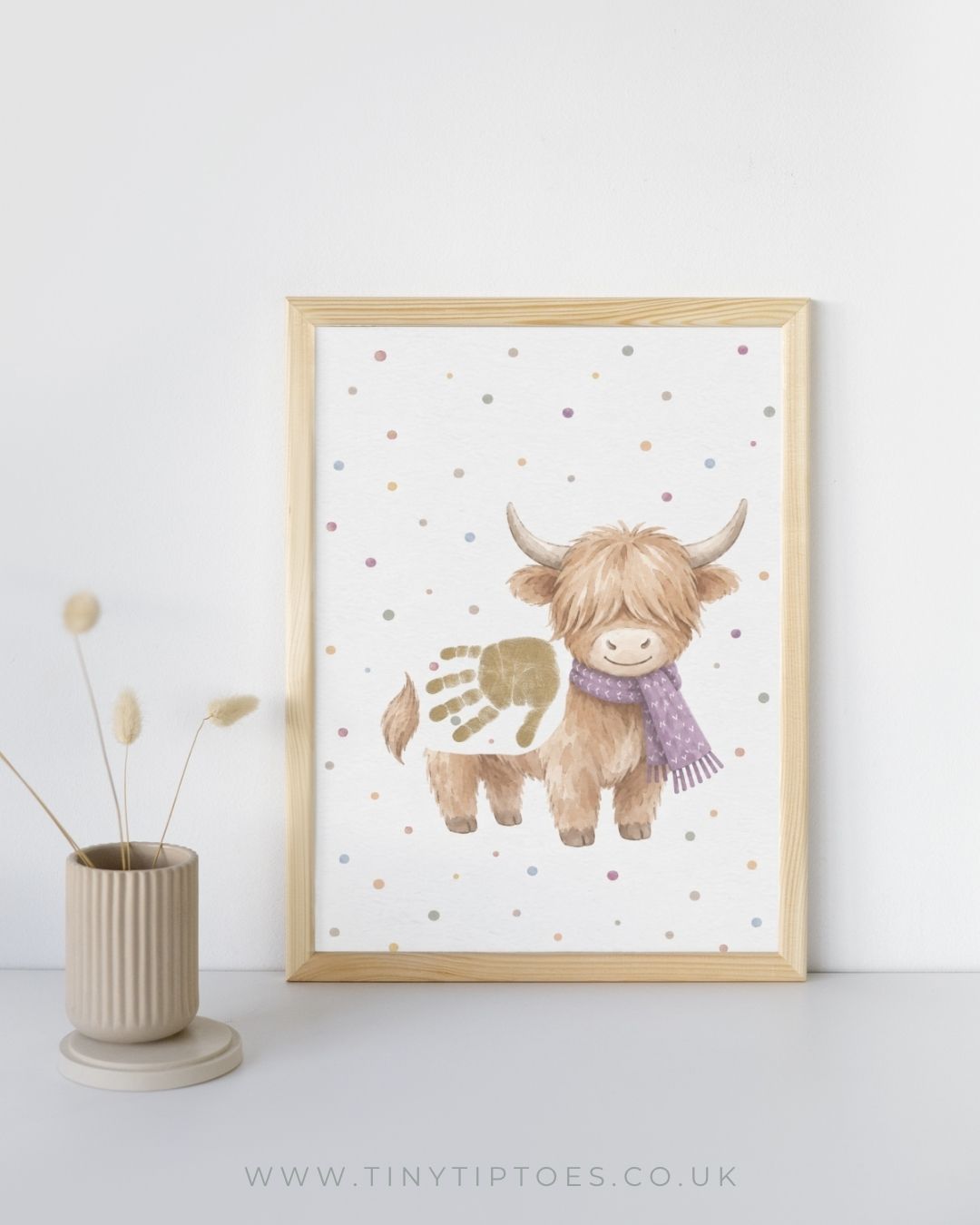 A framed art print of a cute Highland cow with a purple scarf and a gold handprint on its back, styled in a light wood frame next to a beige vase with dried grass on a white background.