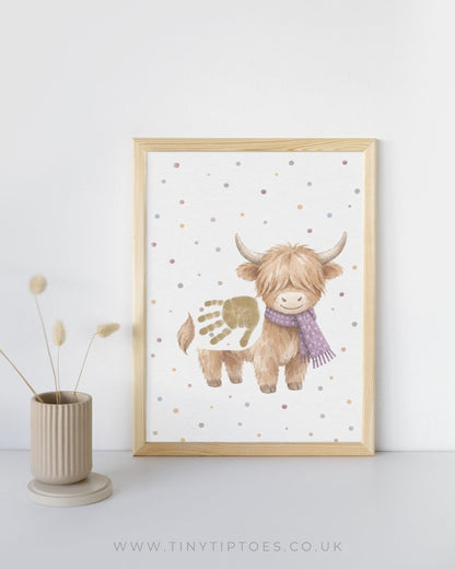 A framed art print of a cute Highland cow with a purple scarf and a gold handprint on its back, styled in a light wood frame next to a beige vase with dried grass on a white background.