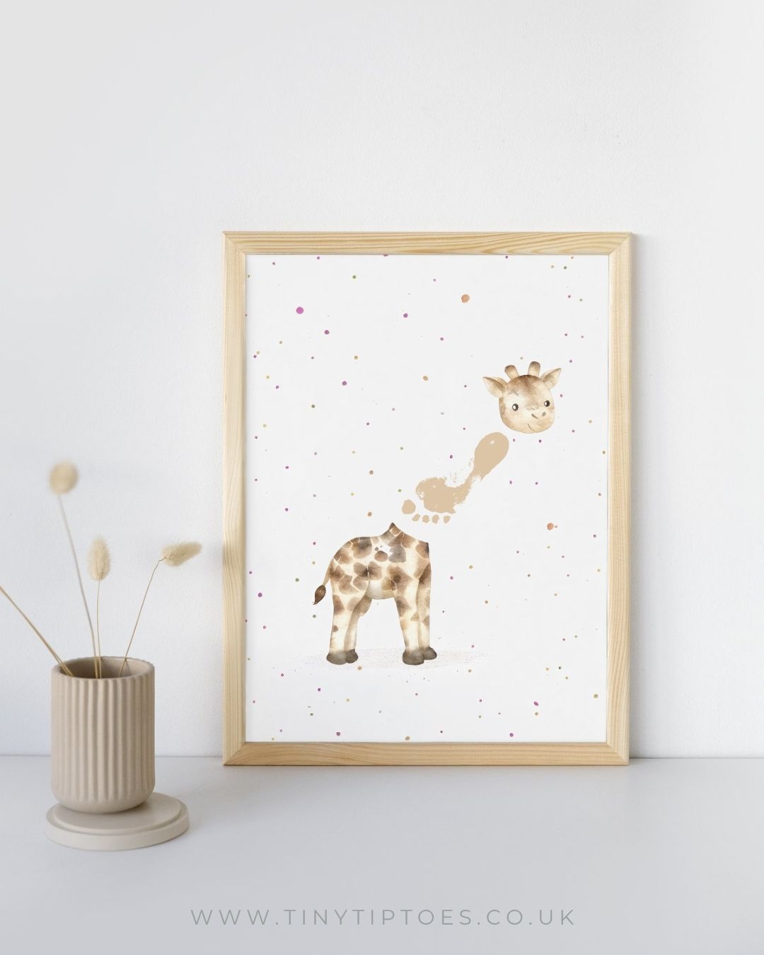Framed artwork of a giraffe with a footprint neck on a white wall with a vase and dried plants.