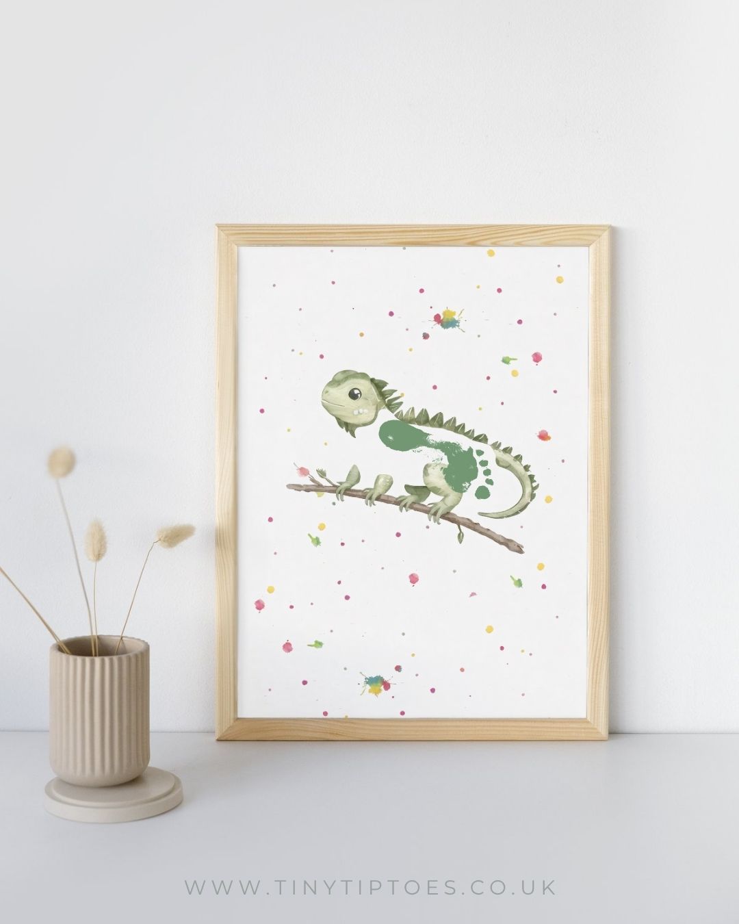 Framed artwork of a green iguana with a footprint bodt on a branch with colorful dots, placed on a white surface with a light background.
