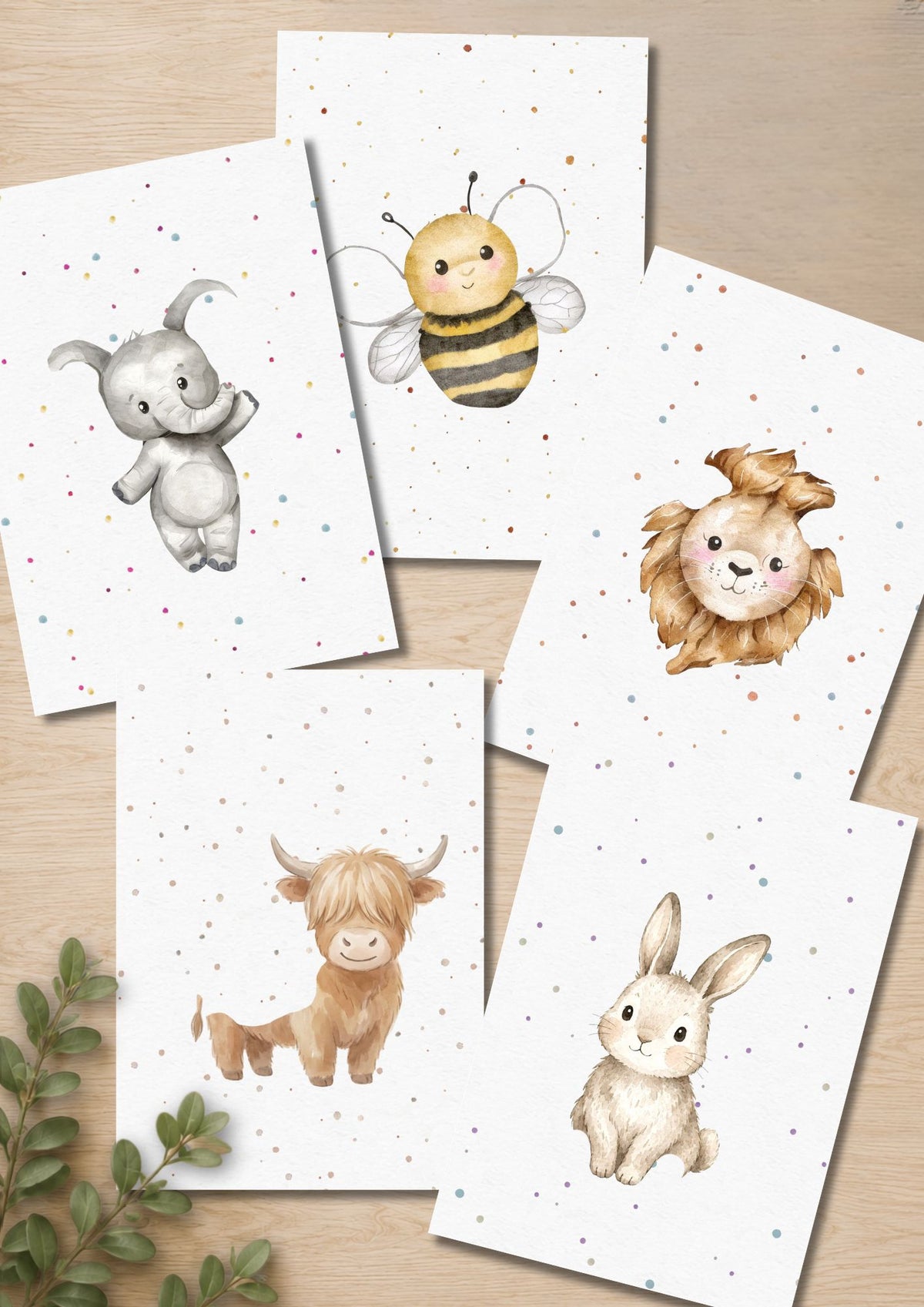 A set of watercolor-style animal art prints arranged on a light wood surface. The prints show a gray elephant, a yellow-and-black bee, a tan lion, a tan Highland cow, and a cream rabbit on white backgrounds with small scattered confetti dots. Animals have missing spaces where baby's hand and footprints will complete the image.