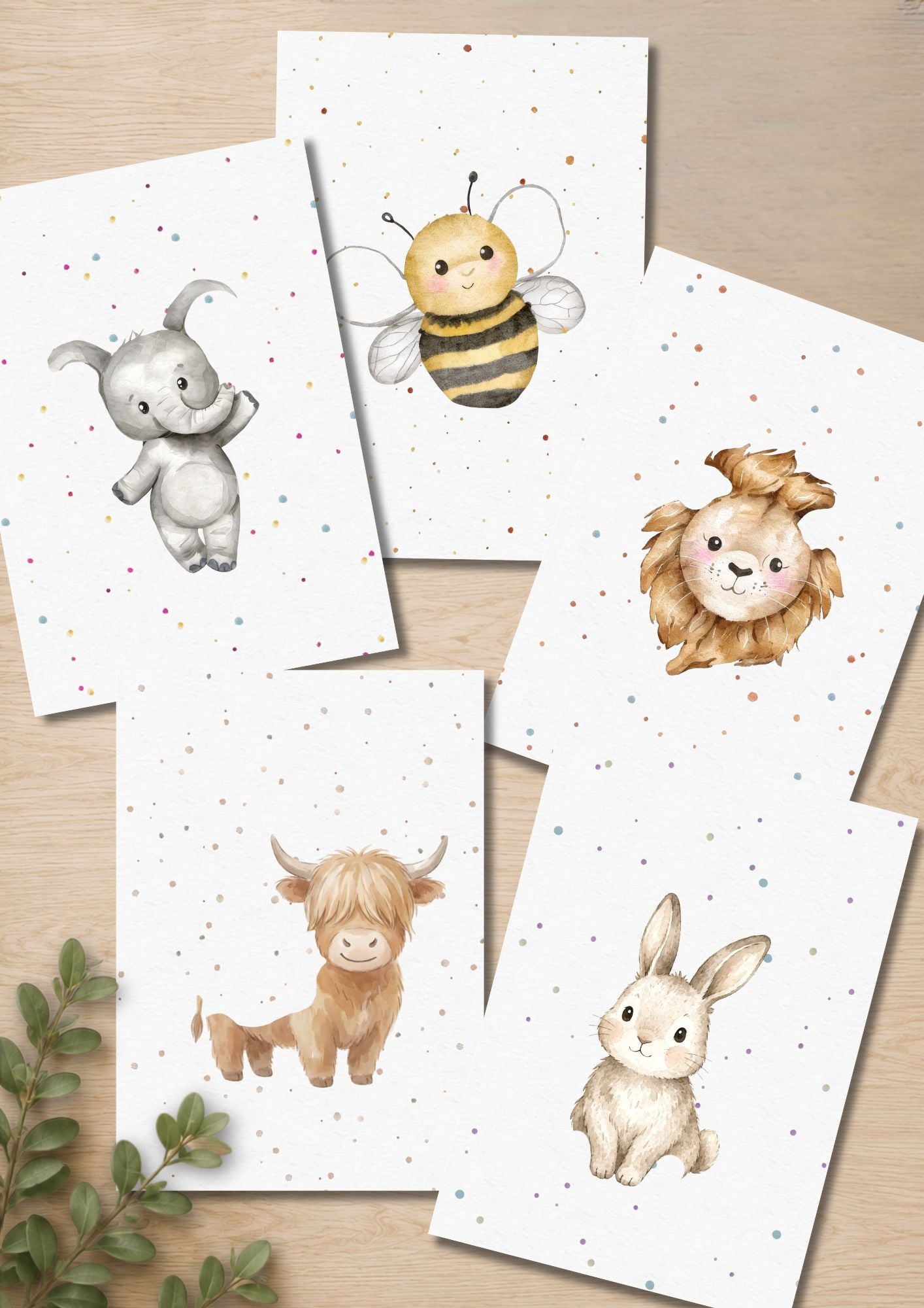 A set of watercolor-style animal art prints arranged on a light wood surface. The prints show a gray elephant, a yellow-and-black bee, a tan lion, a tan Highland cow, and a cream rabbit on white backgrounds with small scattered confetti dots. Animals have missing spaces where baby's hand and footprints will complete the image.