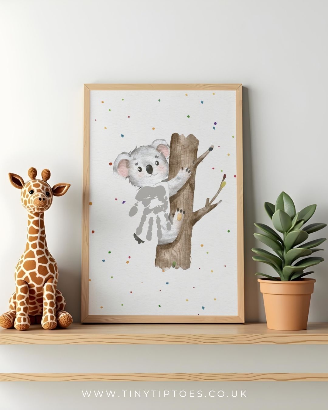 Framed artwork of a koala with handprint body on a tree branch with a toy giraffe and plant on a shelf.