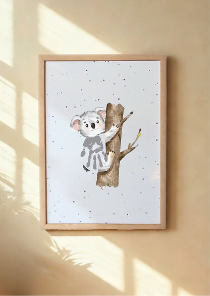 Framed artwork of a koala on a branch with a baby's handprint, on a beige wall.