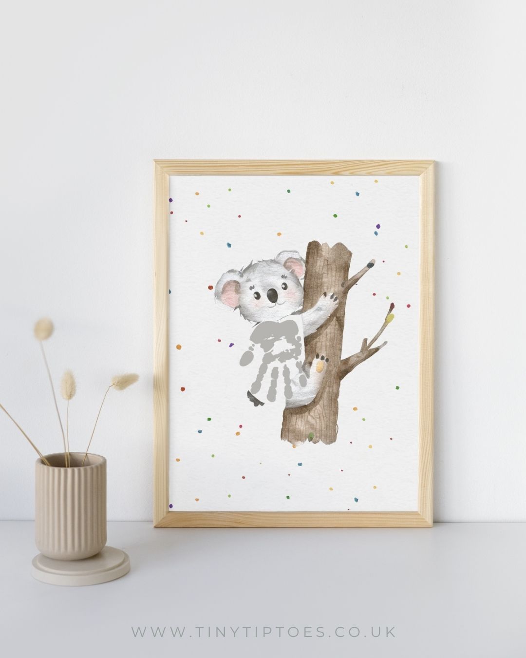 Framed artwork of a koala with a handprint body on a tree branch with colorful dots on a white background, placed on a light surface with a vase.