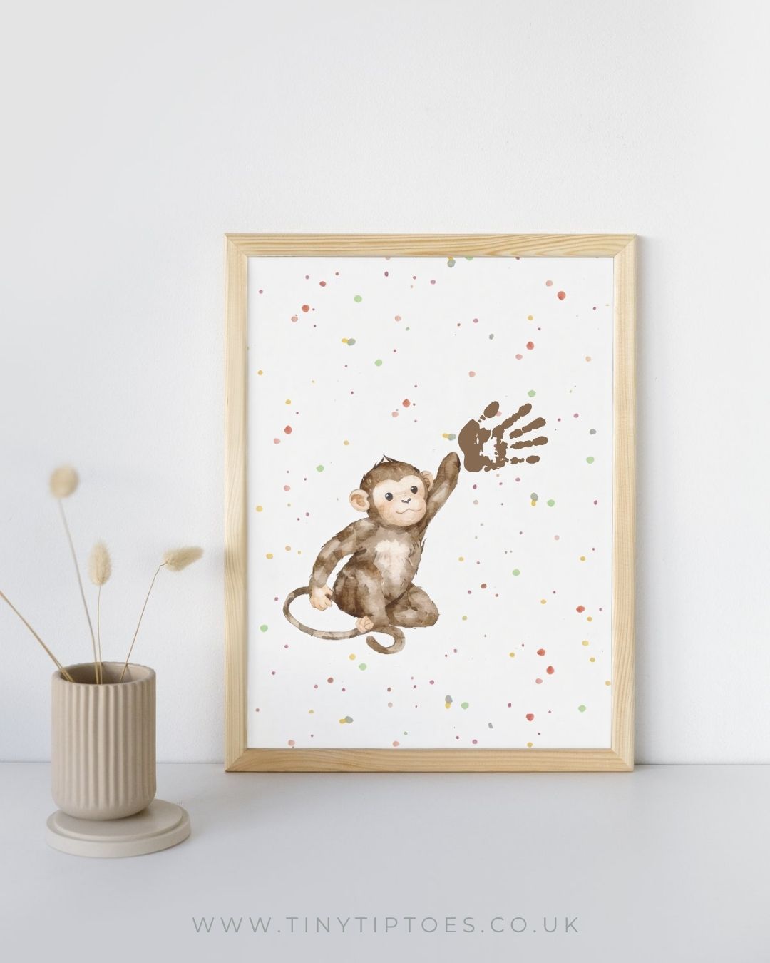 Framed artwork of a monkey with handprint on a white background, displayed on a light gray surface with a vase and dried plants.