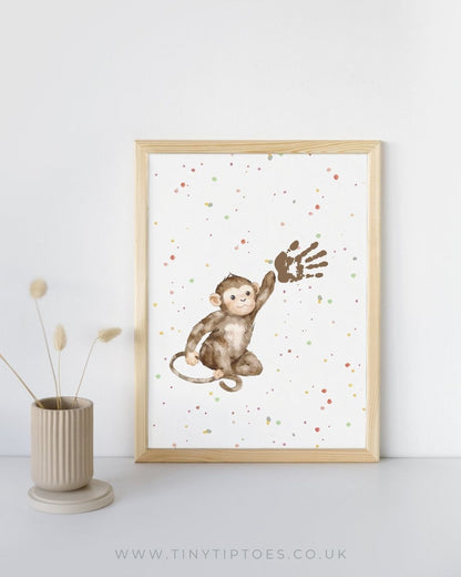 Framed artwork of a monkey with handprint on a white background, displayed on a light gray surface with a vase and dried plants.