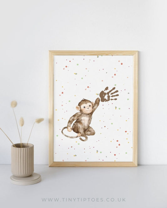 Framed artwork of a monkey with handprint on a white background, displayed on a light gray surface with a vase and dried plants.