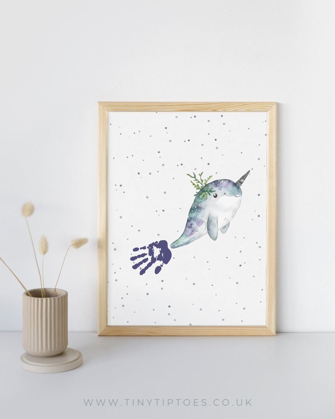 Framed artwork of a narwhal with a handprint tail on a white background, displayed on a light gray surface with a white wall.