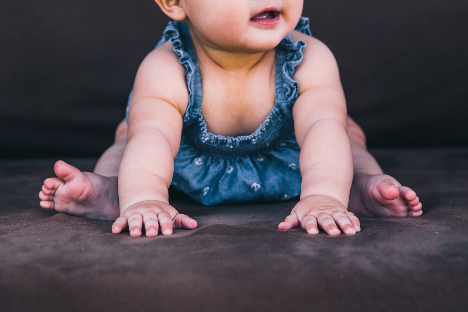 a babt sitting on the loor in a denim dress, arms in front supporting