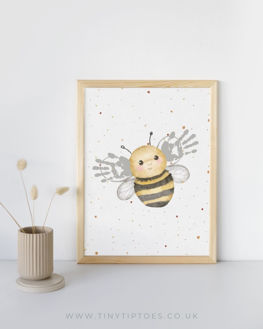Framed artwork of a bee with handprint wings on a white background, next to a vase.